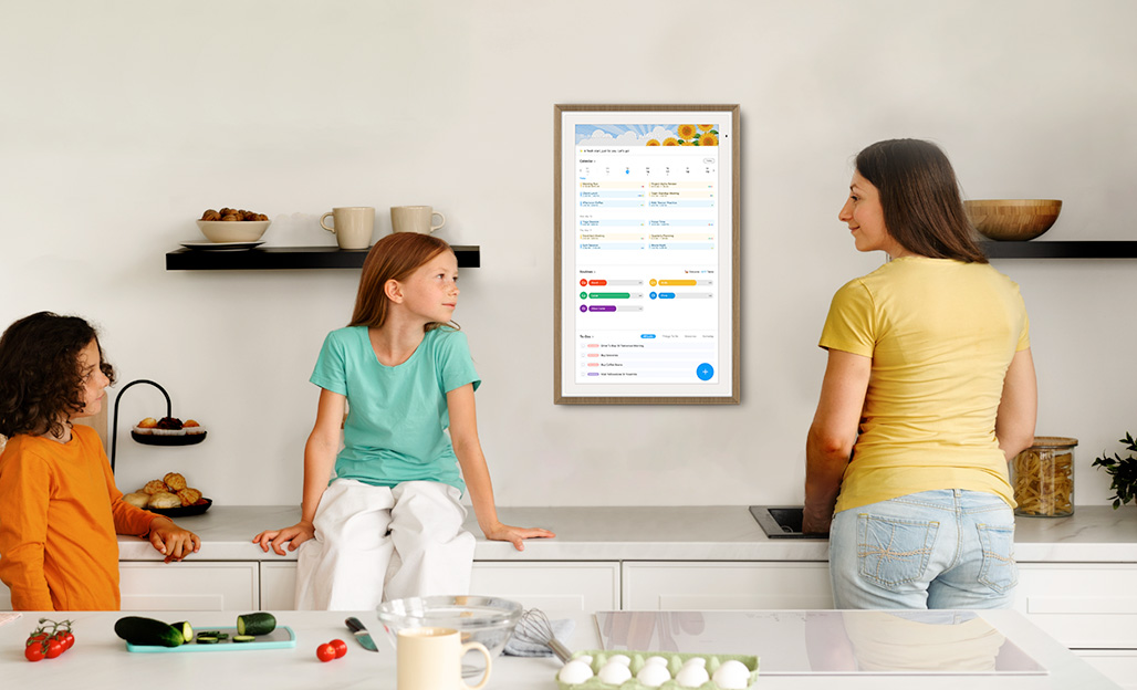 A kitchen scene with a family looking at a wall-mounted digital calendar display. A woman in a yellow shirt and two children are gathered around a kitchen counter with cooking ingredients while viewing the screen that shows a schedule