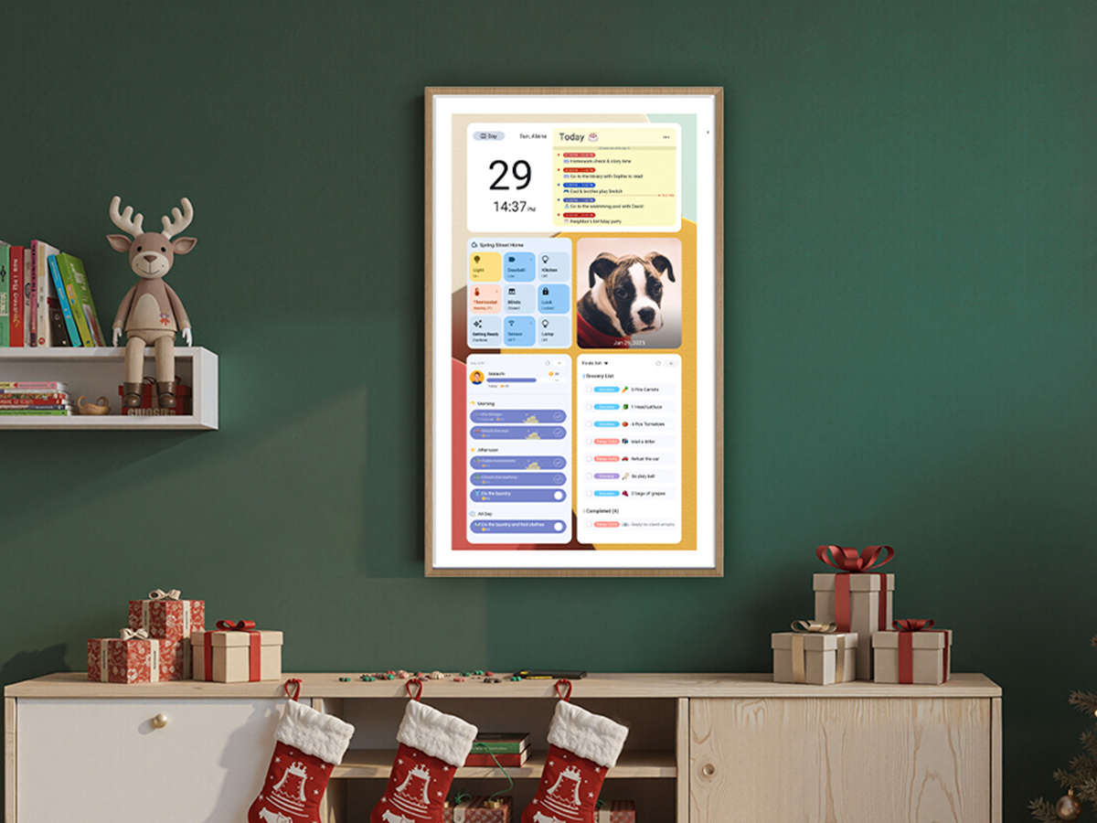 Digital family calendar dashboard mounted in wooden frame on green wall above console table decorated with Christmas stockings and wrapped gifts showing date, weather, pet photo, and task lists