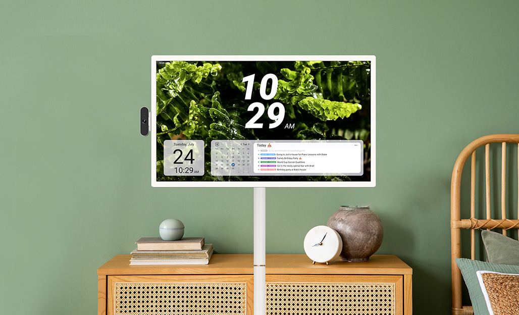 Portable smart TV displaying a digital clock (10:29 AM), calendar, and daily schedule against a green wall, mounted above a wooden cabinet with minimalist decor including books, a small clock, and a ceramic vase