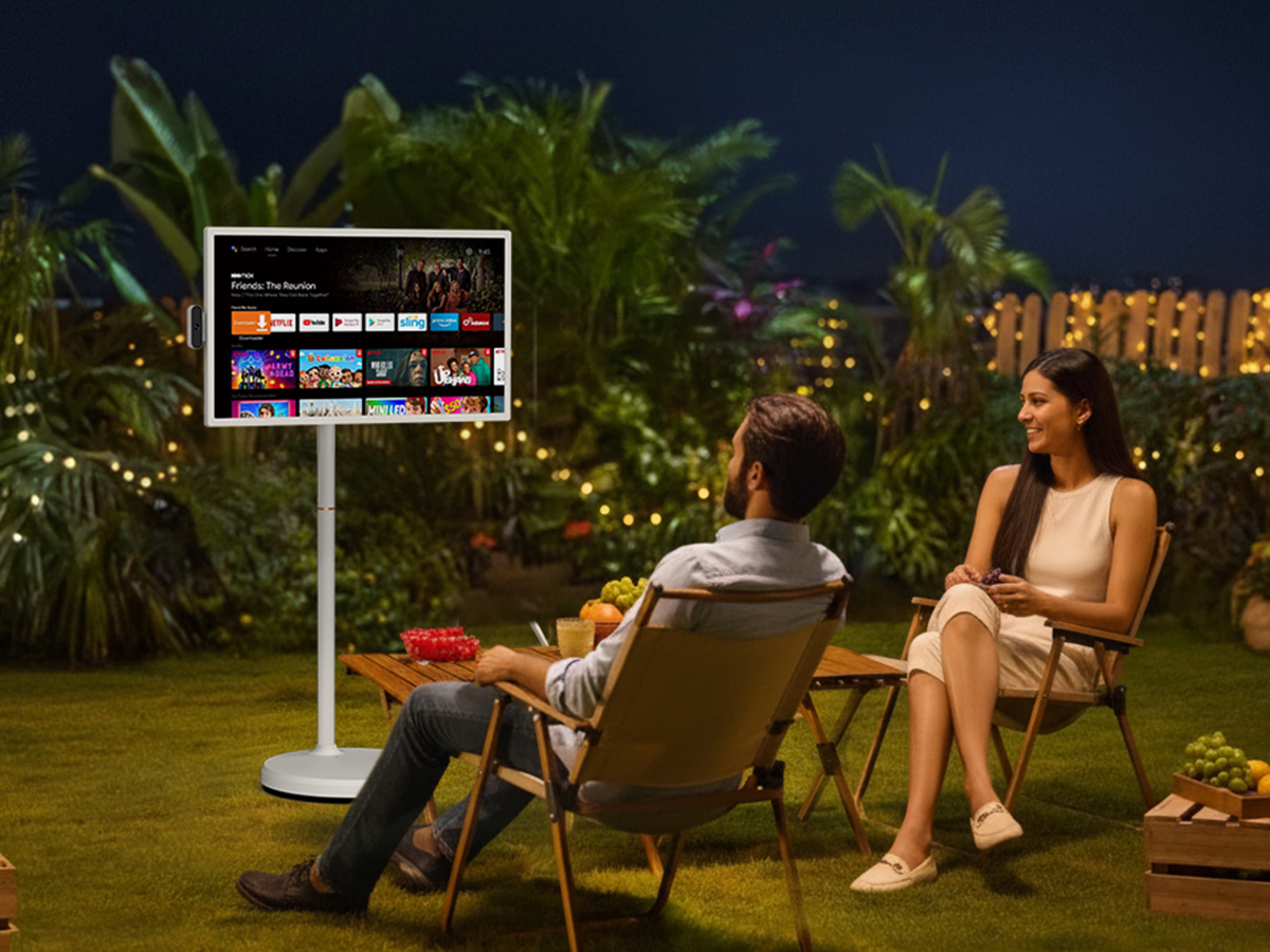 Couple relaxing in wooden chairs on backyard lawn at night watching Friends The Reunion on wall-mounted digital display with streaming service options and string lights in background