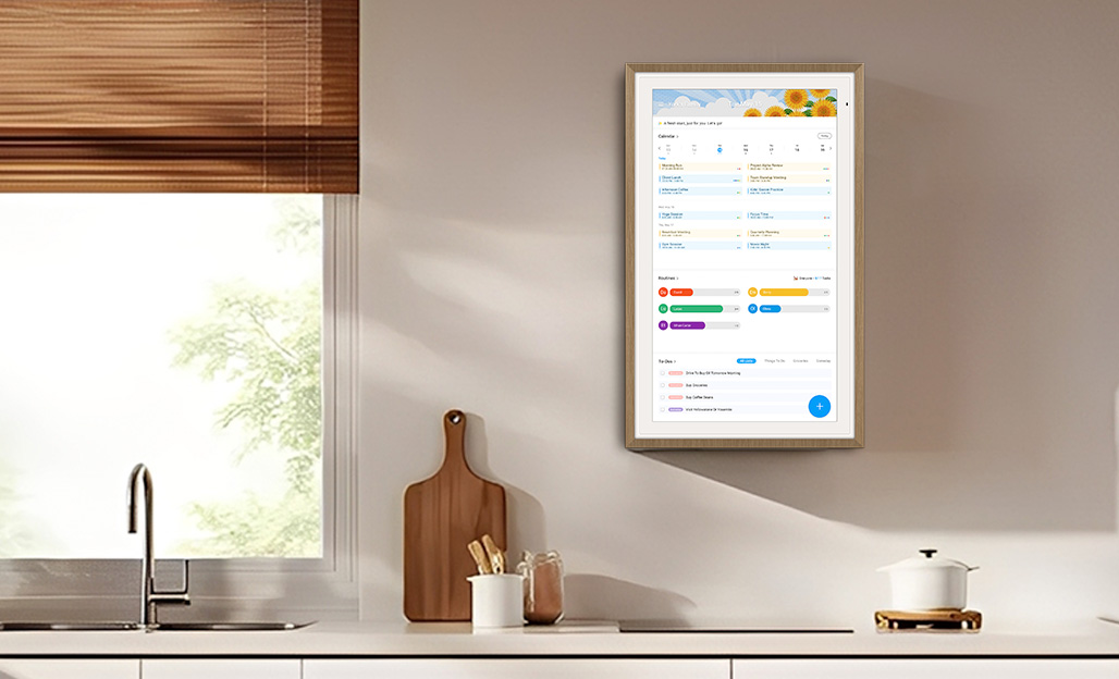 Digital wall calendar mounted in a kitchen setting, displaying a clean interface with color-coded schedule entries, positioned next to a window with wooden blinds and kitchen countertop