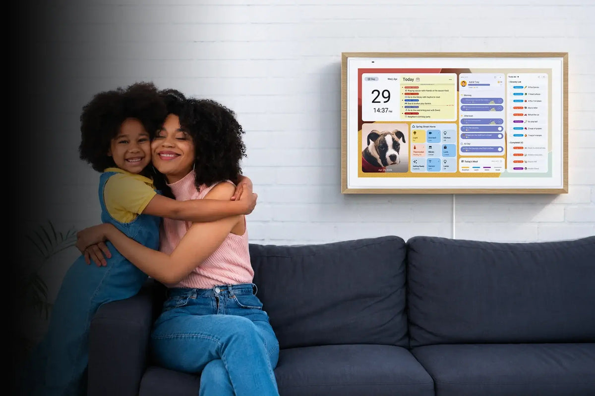 Mother and daughter hugging on sofa, wall‑mount digital calendar displaying organized family schedule dashboard