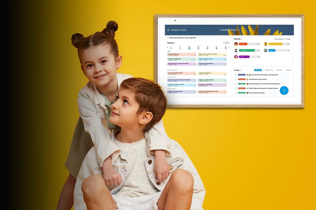 Two children sitting beside a wall-mounted digital calendar displaying a shared family schedule and chores.