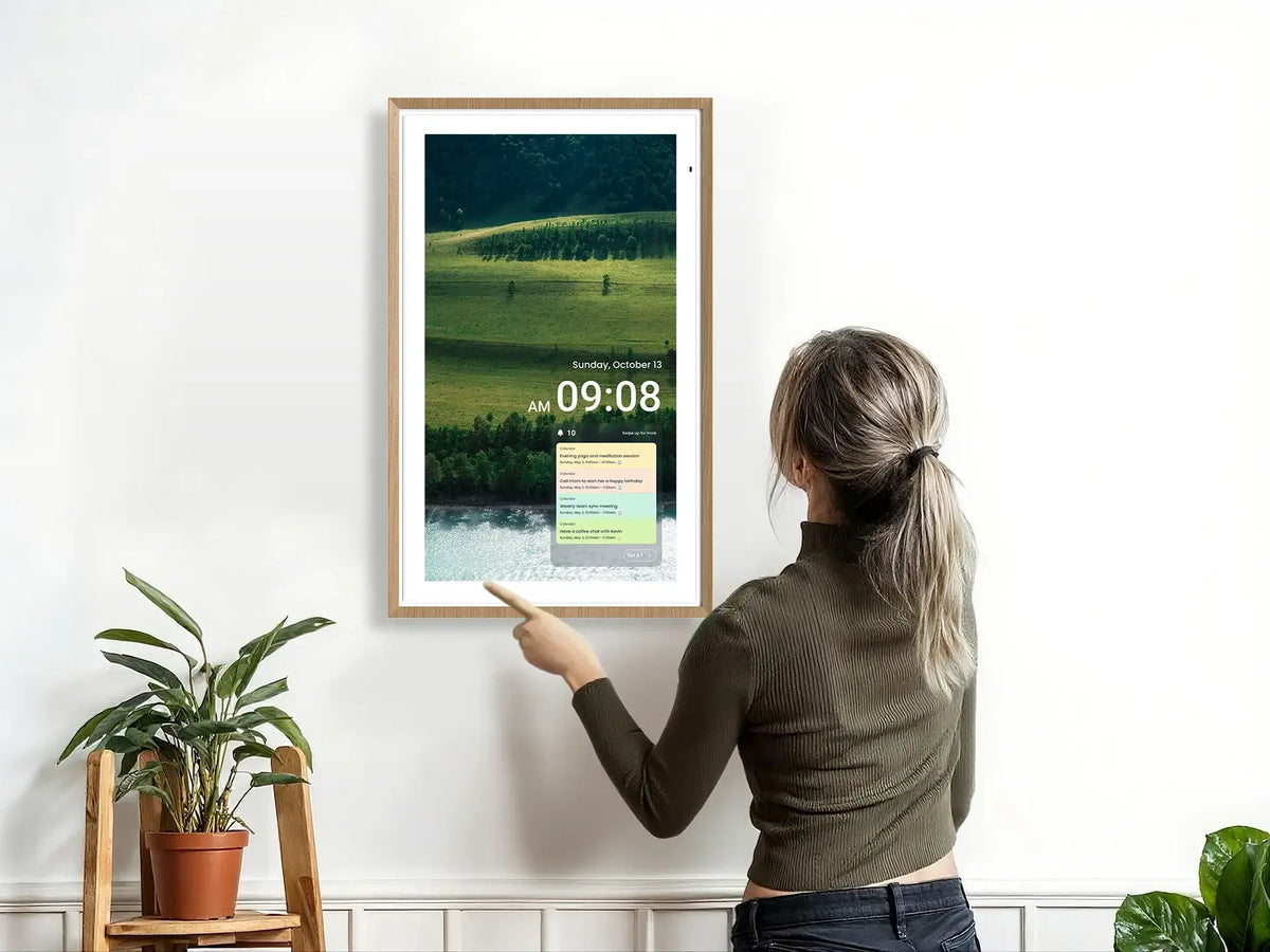 A woman interacting with a wall-mounted Digital Calendar displaying a landscape photo, clock, and daily schedule.